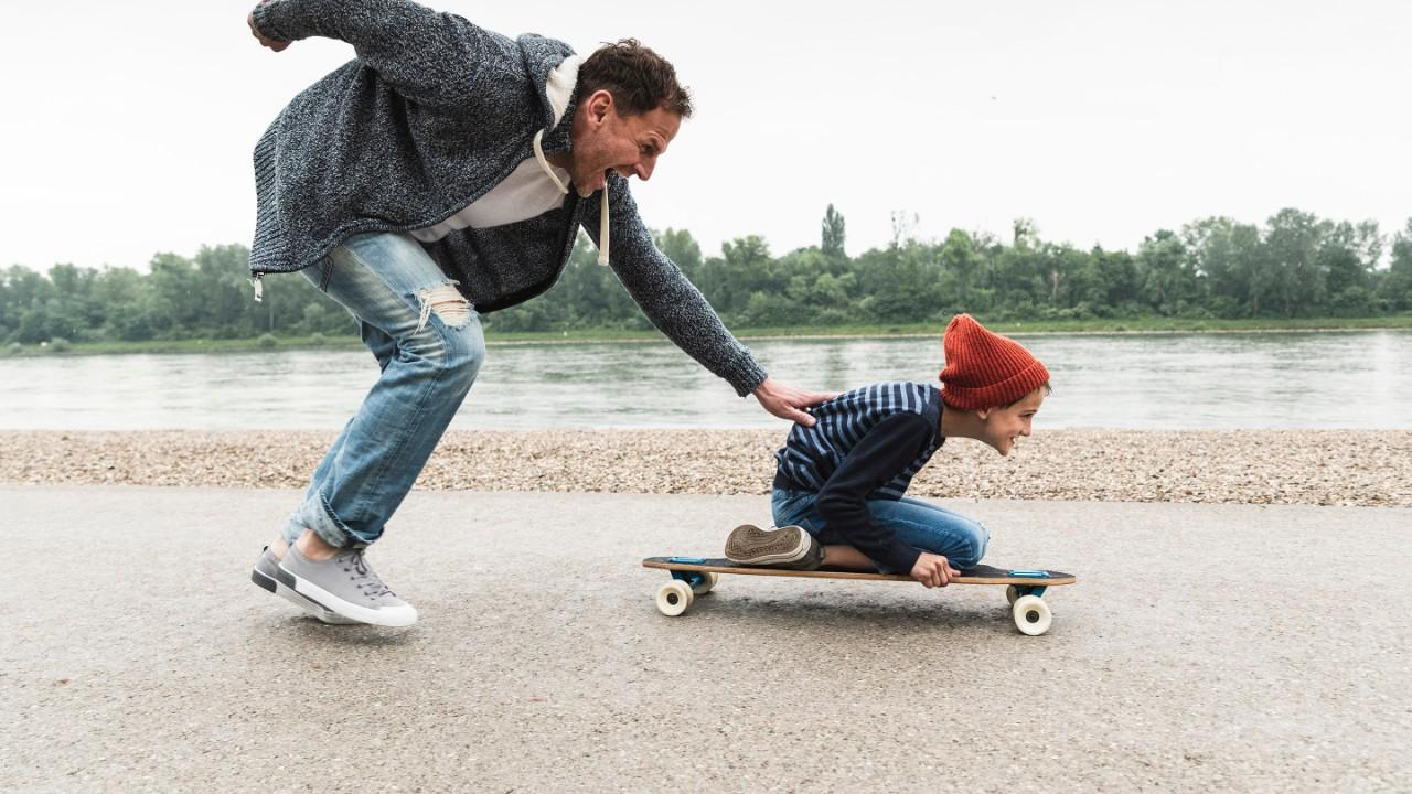 Happy father pushing son on skateboard at the riverside
